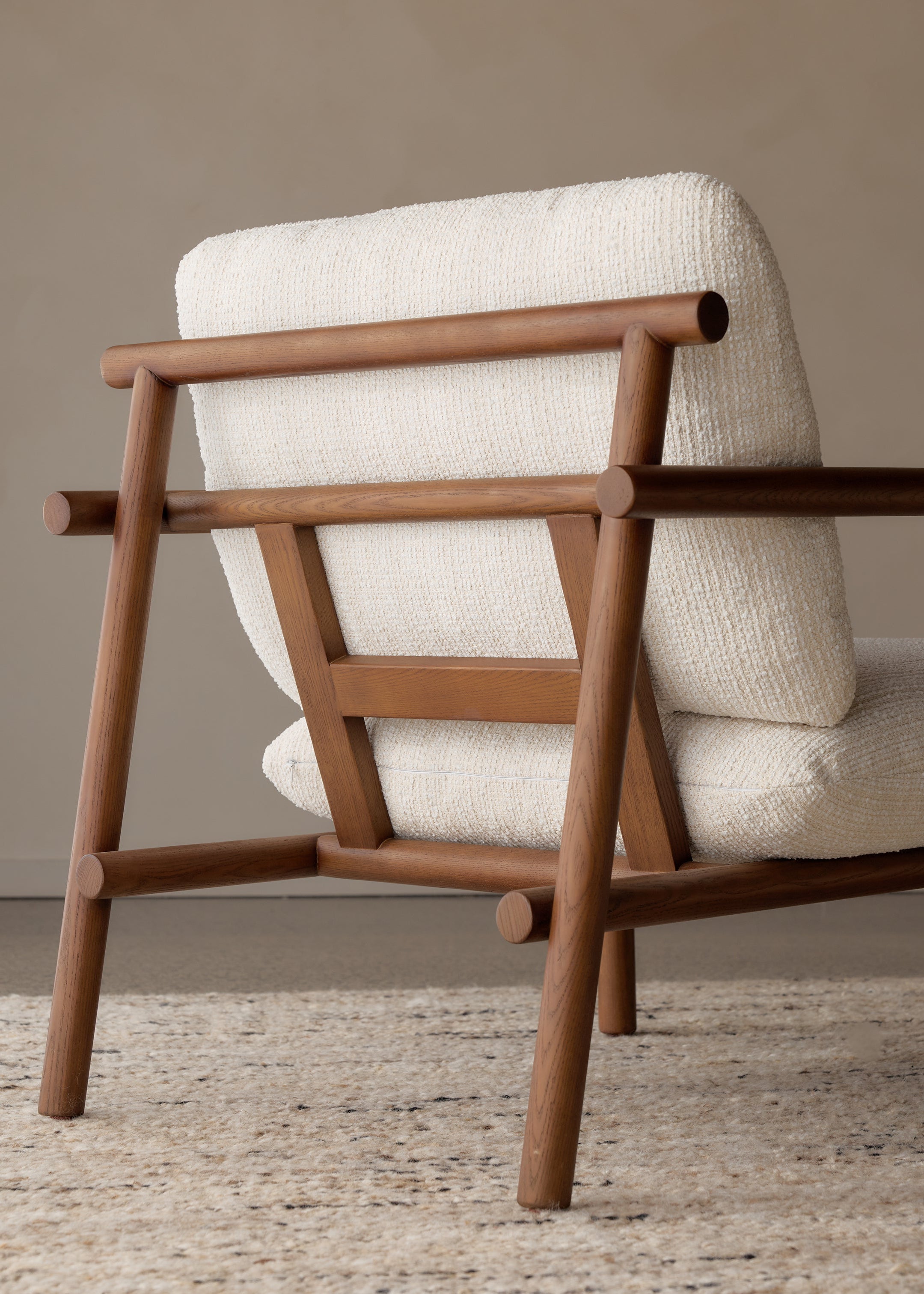 A mid-century modern wooden armchair with a textured off-white bouclé seat and back cushion, shown from a low angle on a neutral rug against a beige wall.